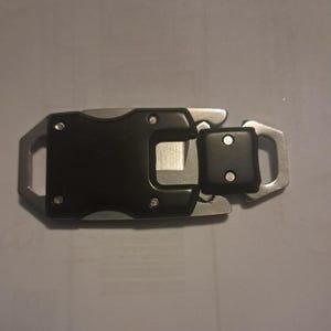 Könnte beinhalten: Eine schwarze und silberne Multi-Tool-Schnalle mit einem Messer und einer quadratischen Komponente. Die Schnalle hat einen rechteckigen schwarzen Mittelteil mit silbernen Metallschlaufen an jedem Ende. Die quadratische Komponente ist ebenfalls schwarz.