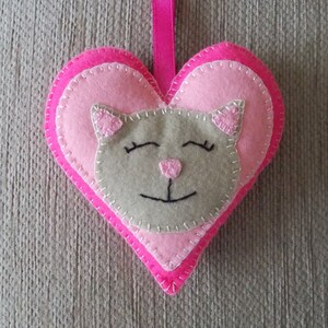 May include: A pink felt heart with a grey felt cat face in the centre. The cat has closed eyes and a small pink nose.