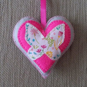 May include: A pink felt heart with a white fabric butterfly with floral print in the centre. The heart has a grey felt border and a pink ribbon.