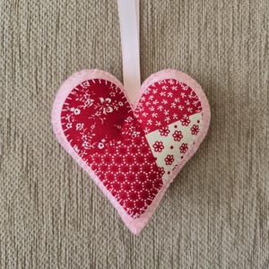 May include: A heart-shaped ornament with a patchwork design. The heart features sections of red fabric with floral patterns and white accents, outlined with pink felt. A white ribbon is attached for hanging.