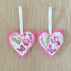 May include: Two heart-shaped decorations with a floral and butterfly design. The ornaments feature a white background with pink flowers and butterflies. Each heart is outlined in pink and has a white ribbon for hanging.