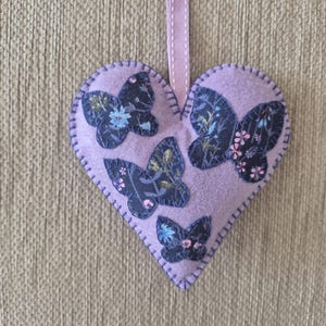 May include: A lilac heart-shaped decoration featuring four butterfly motifs. The butterflies are dark blue with floral patterns in pink, blue, and green. The ornament has a stitched edge and a lilac ribbon for hanging.