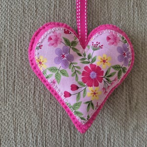 May include: A heart-shaped fabric ornament featuring a light pink background with a floral pattern of red, yellow, and purple flowers with green leaves. The heart is edged with bright pink felt and white stitching, and includes a pink ribbon loop.