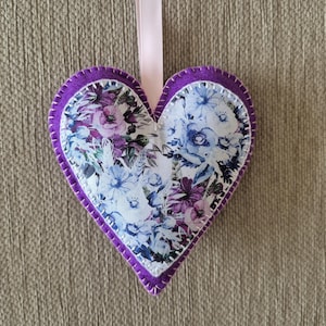 May include: A heart-shaped fabric decoration featuring a floral design in purple, blue, and white. It has a purple border and a pink ribbon. The background is a textured, neutral colour.