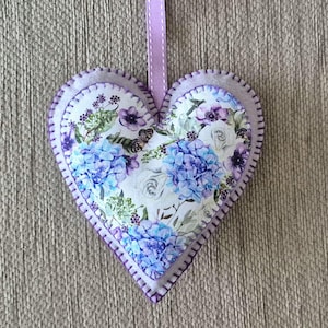 May include: A heart-shaped decoration with a floral design of blue hydrangeas, white roses, and purple details. It has purple stitched edges and a lavender ribbon for hanging. The item is approximately 15 cm in width.