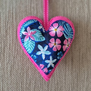 May include: A pink felt heart-shaped ornament with a blue and white floral print. The print features pink flowers, white flowers, and green leaves on a dark blue background.