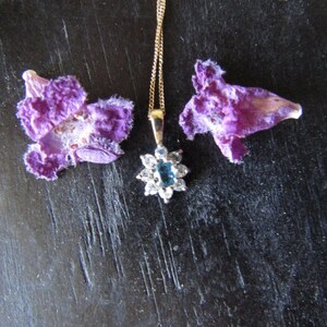 May include: A gold chain necklace with a blue gemstone pendant surrounded by clear stones. The pendant is in the shape of a flower. The necklace is laying on a dark surface with two purple flower petals on either side.