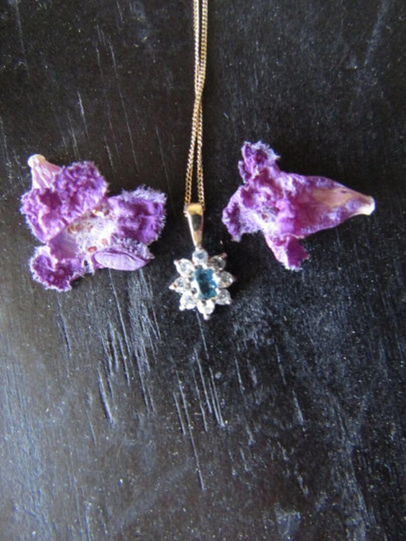 May include: A gold chain necklace with a blue gemstone pendant surrounded by clear stones. The pendant is in the shape of a flower. The necklace is laying on a dark surface with two purple flower petals on either side.