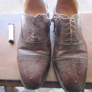 Puede incluir: Un par de zapatos Oxford de cuero marr&oacute;n con cordones. Los zapatos tienen un dise&ntilde;o de brogue y est&aacute;n usados.