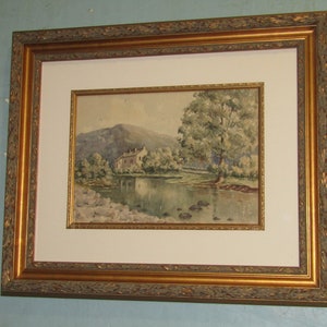 May include: A framed watercolour painting of a landscape scene. The artwork shows a house near a body of water, with trees and a mountain in the background. The painting is set within a gold-coloured ornate frame.