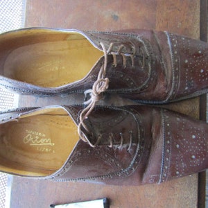 Puede incluir: Un par de zapatos de cuero marr&oacute;n con un dise&ntilde;o de cordones y un patr&oacute;n perforado. Los zapatos tienen una etiqueta que dice "Sapataria Orion".