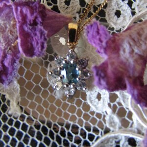 May include: A delicate gold necklace with a blue gemstone pendant surrounded by smaller white gemstones. The necklace is displayed on a white lace background with purple flowers.