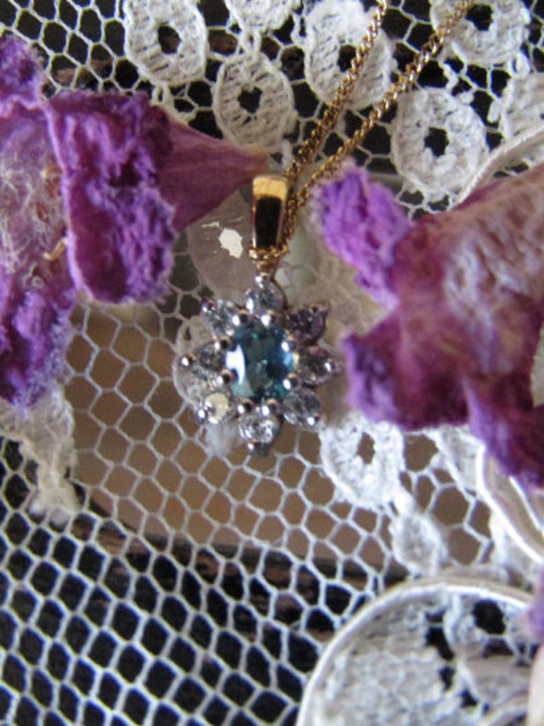 May include: A delicate gold necklace with a blue gemstone pendant surrounded by smaller white gemstones. The necklace is displayed on a white lace background with purple flowers.