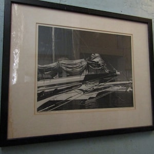 May include: Black and white framed print of a sailboat. The boat is docked, with its sails furled and covered. Ropes and rigging are visible, and the image is set against a dark background.