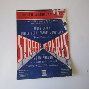 May include: A vintage sheet music cover for the song "Streets of Paris" featuring a red, white and blue colour scheme. The cover features a title card with the song title in bold red letters and the names of the performers, Bobby Clark, Luella Gear, Abbott & Costello, and Jean Sablon, in white letters. The cover also includes a depiction of streetlights and a pavement.