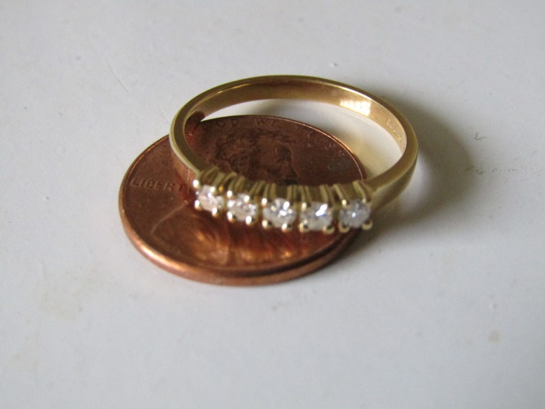 May include: A gold band ring with five small diamonds set in a row. The ring is placed on top of a US penny.