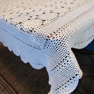 May include: White crocheted tablecloth with a delicate floral pattern. The tablecloth is draped over a wooden table and has a scalloped edge.