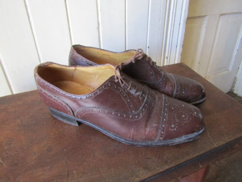 Puede incluir: Un par de zapatos de cuero marr&oacute;n con detalles de punta de ala. Los zapatos tienen un cierre de cordones y un acabado pulido.
