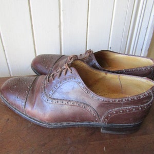 Puede incluir: Un par de zapatos Oxford de cuero marr&oacute;n con detalles de brogue. Los zapatos tienen un cierre de cordones y una punta redonda.