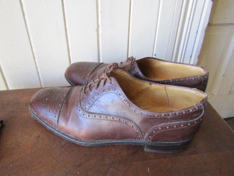 Puede incluir: Un par de zapatos Oxford de cuero marr&oacute;n con detalles de brogue. Los zapatos tienen un cierre de cordones y una punta redonda.