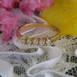 May include: A gold band ring with a row of small, clear diamonds set in the band. The ring is on a white lace background.