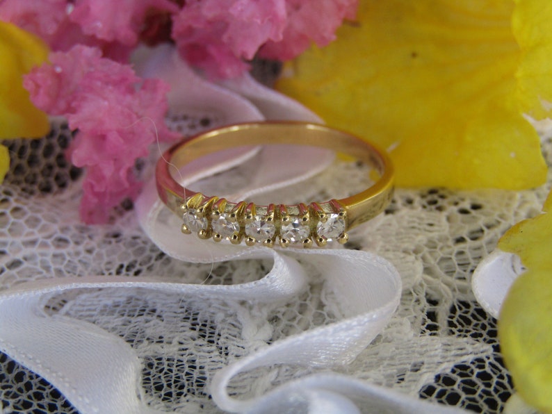 May include: A gold band ring with a row of small, clear diamonds set in the band. The ring is on a white lace background.