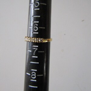 May include: A gold band ring with a row of small clear stones. The ring is sized on a ruler that shows the ring is size 7.
