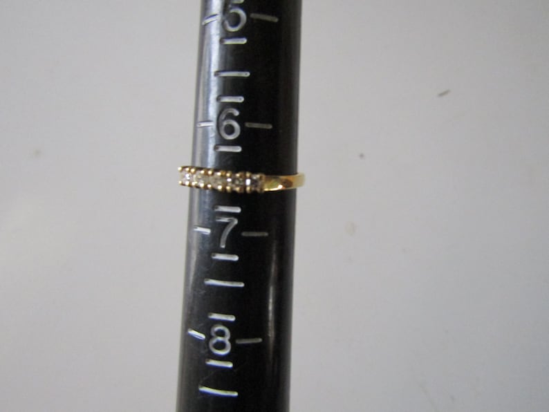 May include: A gold band ring with a row of small clear stones. The ring is sized on a ruler that shows the ring is size 7.