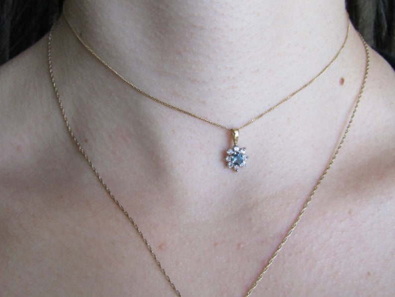 May include: A gold chain necklace with a delicate pendant featuring a blue gemstone surrounded by smaller clear gemstones.