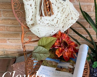 May include: A white crocheted tote bag with a brown and orange tassel hanging from the handle. The bag is sitting on a rusty metal chair with a magazine and a fake sunflower on top.