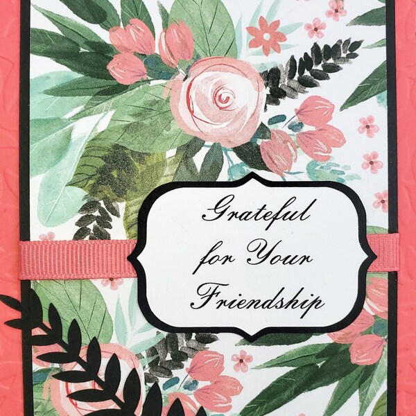 Puede incluir: Una tarjeta floral rosa y verde con un borde negro y una cinta negra. La tarjeta tiene una etiqueta blanca con el texto "Grateful for Your Friendship".
