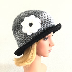 Puede incluir: Un gorro de punto gris y negro con una flor blanca de ganchillo en el lateral. El gorro tiene un borde negro.