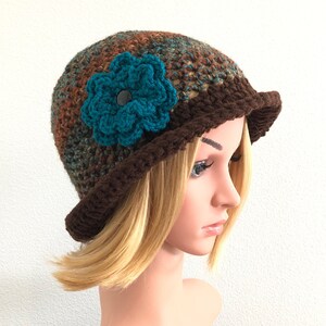 Puede incluir: Un gorro de punto marr&oacute;n con un adorno de flor turquesa. El gorro tiene un ala ancha y est&aacute; hecho con un hilo texturizado.