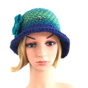 Puede incluir: Un sombrero de crochet azul y verde con un adorno de flor. El sombrero tiene un ala ancha y est&aacute; hecho con un punto texturizado.