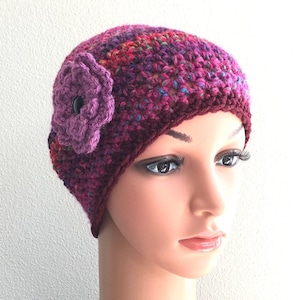May include: A crocheted beanie hat in shades of purple, pink, and red with a large purple crocheted flower on the side.