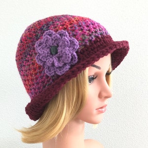 Puede incluir: Un gorro de crochet con un adorno de flor morada. El gorro est&aacute; hecho de un hilo multicolor con tonos de rojo, rosa y morado. El borde del gorro es de color burdeos s&oacute;lido.