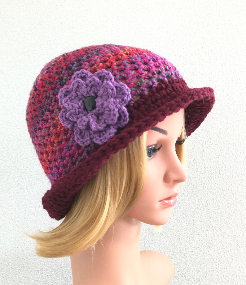 Puede incluir: Un gorro de crochet con un adorno de flor morada. El gorro est&aacute; hecho de un hilo multicolor con tonos de rojo, rosa y morado. El borde del gorro es de color burdeos s&oacute;lido.