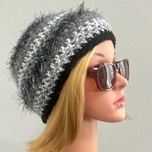 May include: A handmade crochet beanie in shades of gray, white, and black. The hat has a textured, fuzzy appearance and a black brim. The beanie is designed to be worn, offering warmth and style. A fashionable accessory for cold weather.