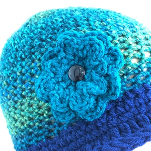 Puede incluir: Un gorro de punto azul con una gran flor turquesa y un bot&oacute;n negro en el centro.
