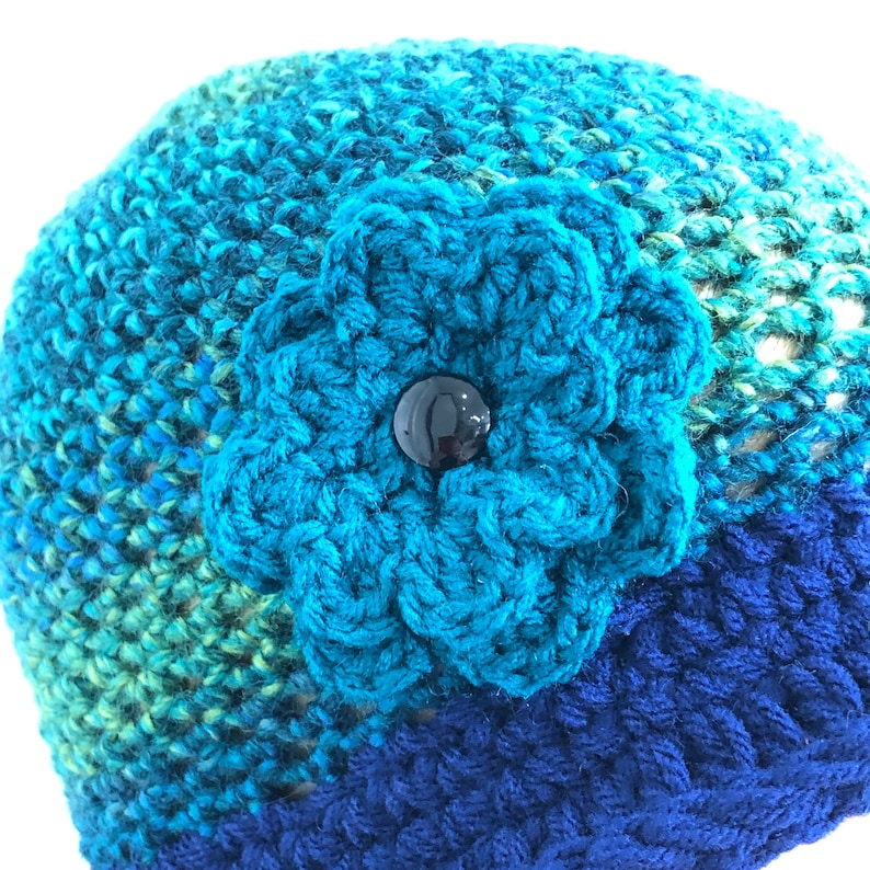 Puede incluir: Un gorro de punto azul con una gran flor turquesa y un bot&oacute;n negro en el centro.