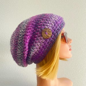 May include: A crocheted beanie in shades of purple, magenta, and grey, featuring a textured yarn pattern and a single round, brown wooden button attached to the side. The hat has a slouchy fit and is crafted with a visible, thick stitch design.