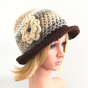 Puede incluir: Un gorro de punto marr&oacute;n y beige con un borde marr&oacute;n y una flor de ganchillo beige en un lado.