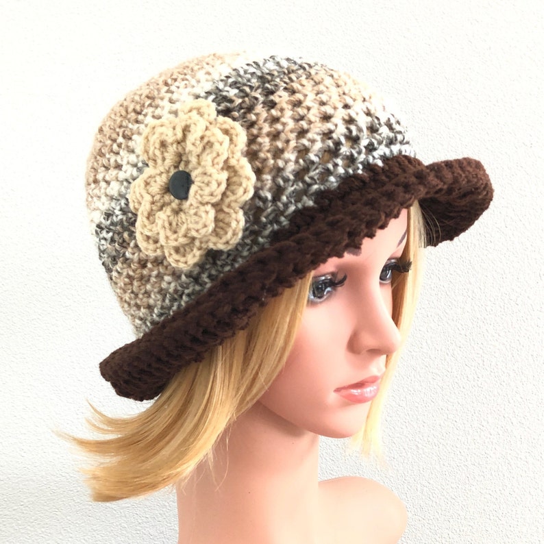 Puede incluir: Un gorro de punto marr&oacute;n y beige con un borde marr&oacute;n y una flor de ganchillo beige en un lado.