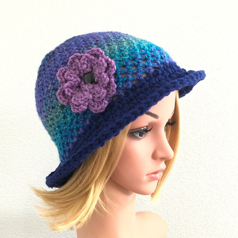 Puede incluir: Un gorro de punto azul y morado con un adorno de flor morada. El gorro tiene un ala ancha y est&aacute; hecho con un punto texturizado.