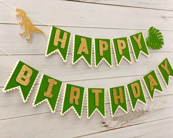 May include: A two-tiered birthday banner featuring green pennants with white zigzag edges and gold glitter letters spelling out HAPPY BIRTHDAY. The banner includes a gold glitter dinosaur cutout on the left and a green tropical leaf on the right.