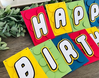 May include: A colorful paper banner featuring the text HAPPY BIRTHDAY, with each letter mounted on a rectangular cardstock base in red, yellow, green, or blue. Each card has raised circular accents and white letters with black outlines.