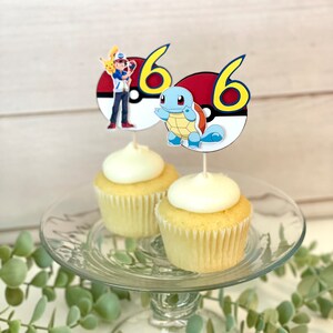 K&ouml;nnte beinhalten: Zwei Vanille-Cupcakes mit wei&szlig;em Zuckerguss, jeweils mit einem runden Papier-Topper versehen. Ein Topper zeigt Ash Ketchum mit Pikachu und die Zahl 6, der andere Schiggy und die Zahl 6, beide vor einem Pok&eacute;ball-Motiv als Hintergrund.