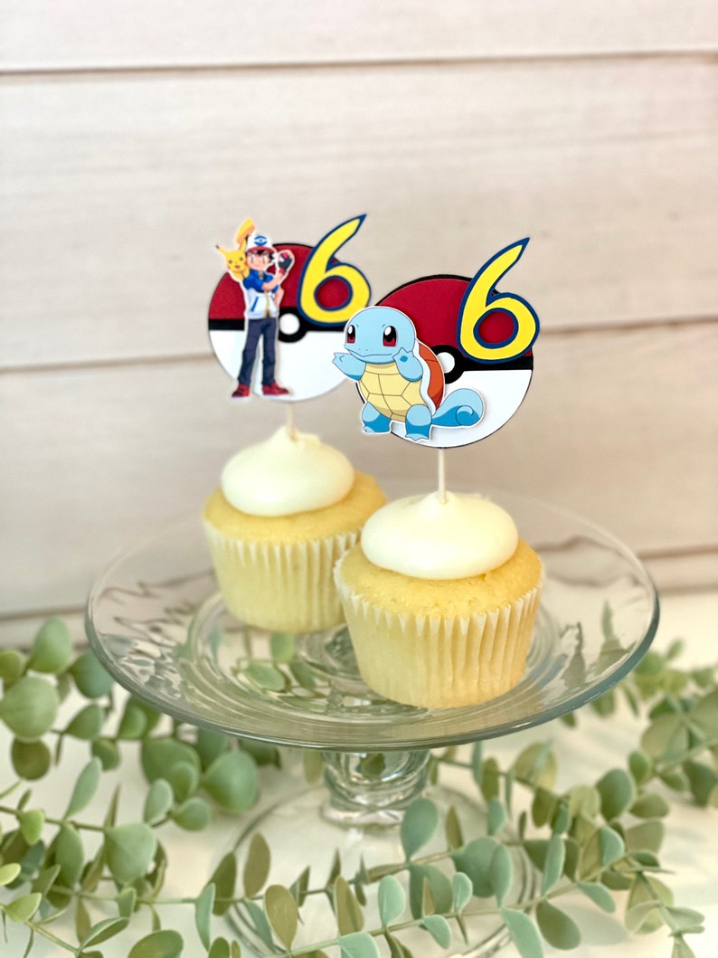 K&ouml;nnte beinhalten: Zwei Vanille-Cupcakes mit wei&szlig;em Zuckerguss, jeweils mit einem runden Papier-Topper versehen. Ein Topper zeigt Ash Ketchum mit Pikachu und die Zahl 6, der andere Schiggy und die Zahl 6, beide vor einem Pok&eacute;ball-Motiv als Hintergrund.