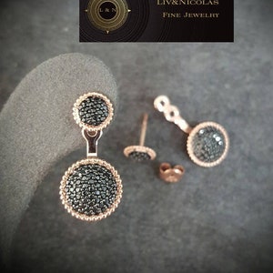 May include: A pair of rose gold earrings with black stones. The earrings are designed with two round stones, one larger than the other, connected by a small chain. The earrings are shown on a gray surface.