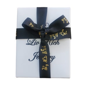 May include: A white gift box with a black ribbon tied in a bow. The ribbon has gold lettering that says "Liv Rich Jewelry" and "By Liv Rich".